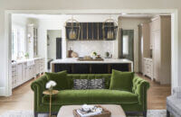 A Southern moern farmhouse kitchen with soft, off-white cabinets, a black metal hood, dark gray kitchen island, and olive green accents including the green couch in the neighboring living room.